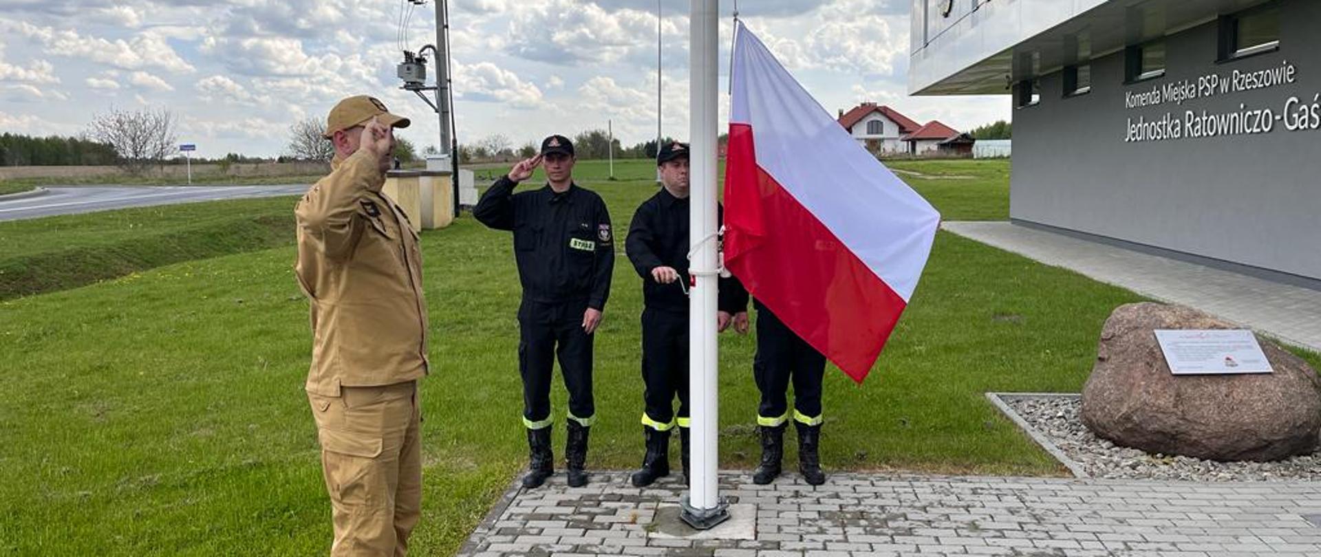 Zdjęcie przedstawia czterech strażaków stojących przy maszcie. Dwóch z nich salutuje, natomiast jeden przy użyciu korby podnosi Flagę Państwową na maszt. W tle widoczny jest budynek Jednostki Ratowniczo-Gaśniczej nr 3 w Rzeszowie.