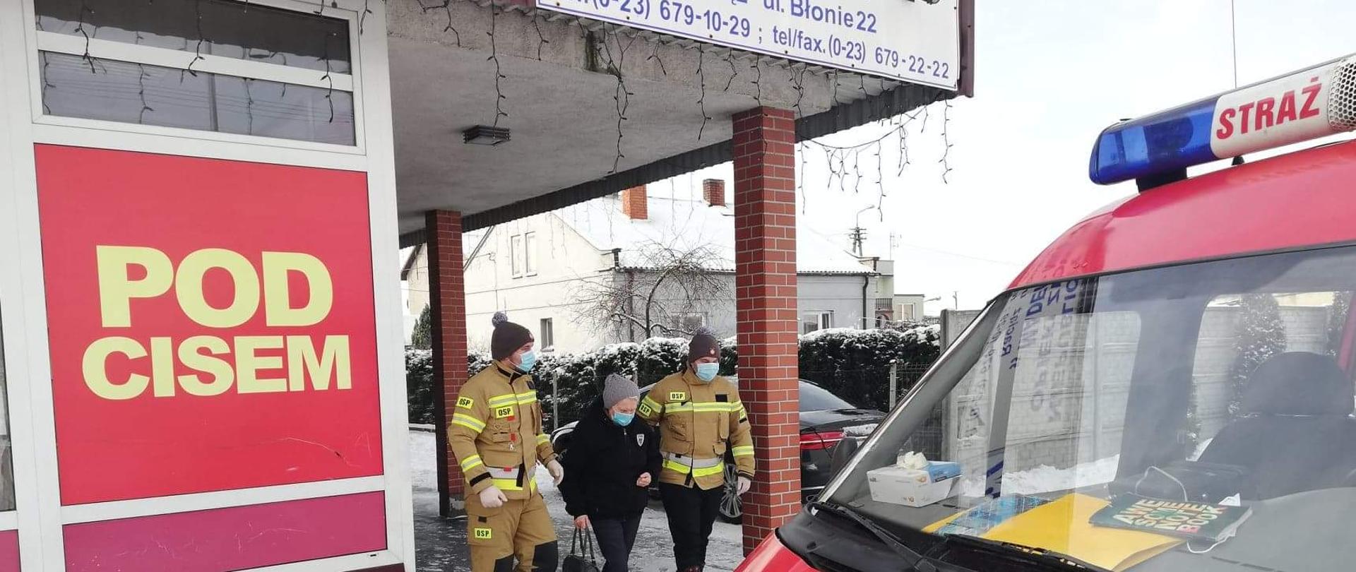 Dwóch strażaków ochotniczej straży pożarnej, ubranych w żółte stroje specjalne w maseczkach wychodzących z budynku przychodni prowadzi do czerwonego samochodu stojącego przed budynkiem starszą kobietę.