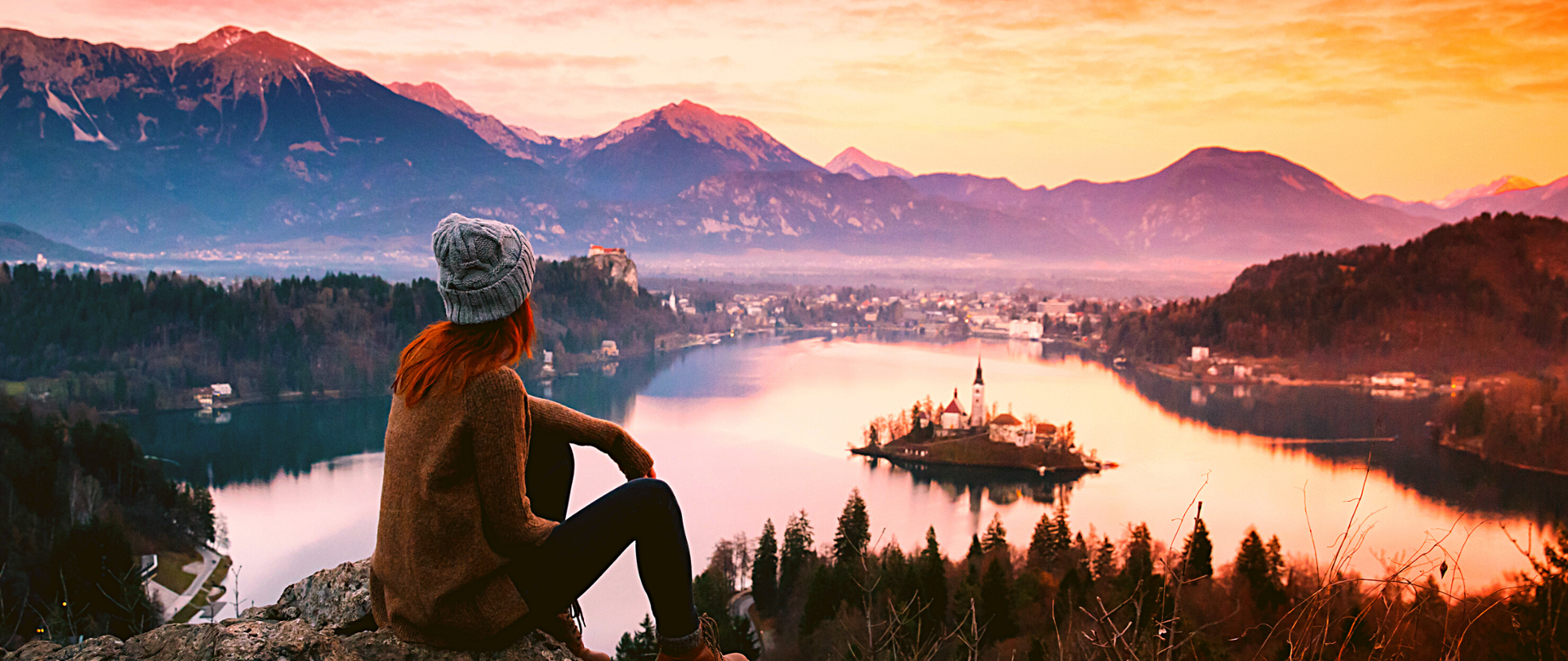 Kobieta w wełnianej czapce siedząca na wzgórzu wpatrzona w krajobraz poniżej - jezioro Bohinjsko z kościołem na wyspie