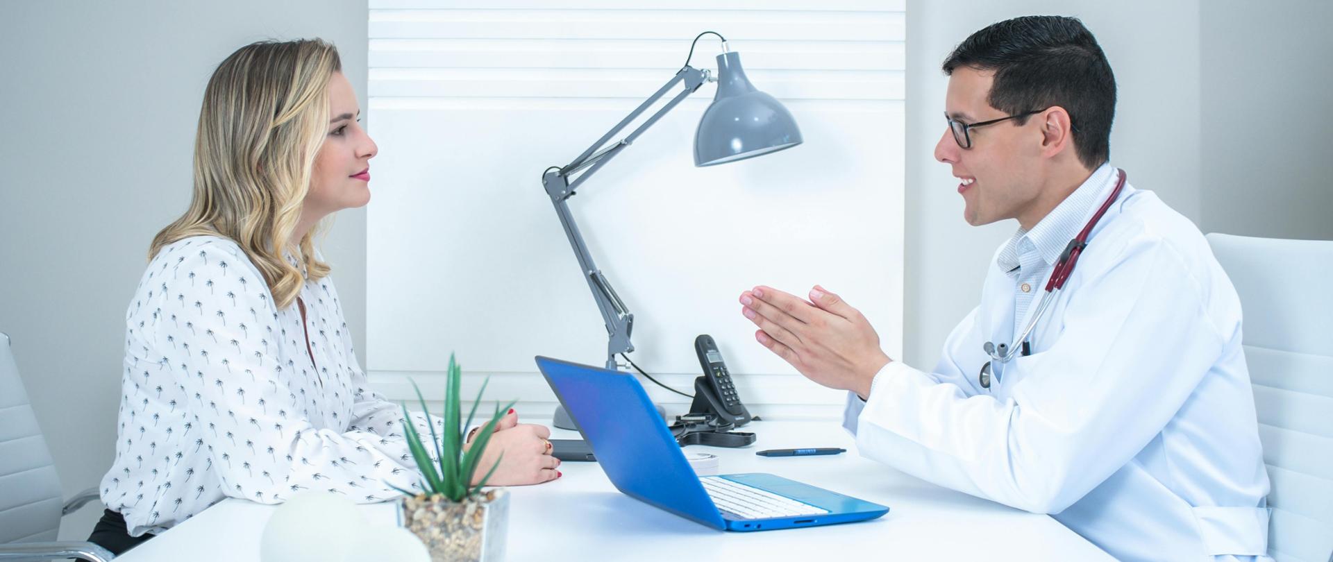 Kobieta słucha lekarza, oboje siedzą przy białym biurku. Na stole leży niebieski laptop, lampa i telefon.
