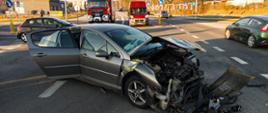 Rozbity przód auta osobowego koloru szarego. W tle samochód strażacki oraz inne pojazdy przejeżdżające przez skrzyżowanie. Pora dzienna, słoneczna.