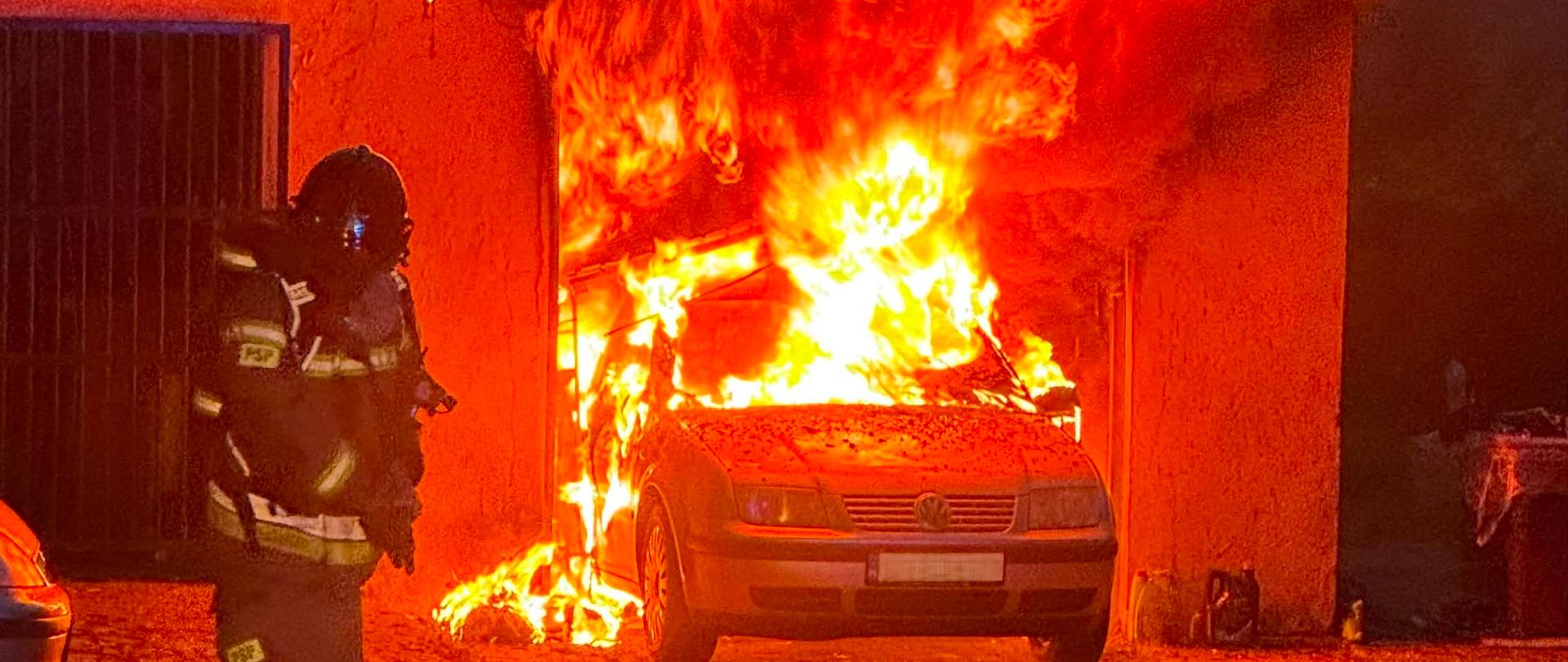 Duży pożar samochodu w garażu z silnymi, wysokimi płomieniami buchającymi z wnętrza budynku. Na pierwszym planie po lewej stronie stoi strażak w pełnym rynsztunku bojowym.