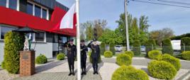 Zdjęcie zrobione w dzień przed budynkiem JRG PSP Sandomierz. Na zdjęciu widać strażaków trzech stojących przed masztem flagowym. Jeden z nich wciąga flagę państwową na maszt. 