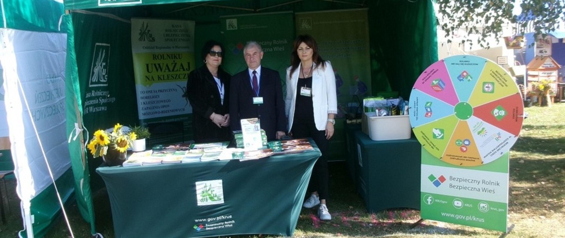 Stoisko informacyjno-prewencyjne Kasy z materiałami informacyjnymi KRUS, wewnątrz stoiska pozują do zdjęcia dwie kobiety i mężczyzna. Po prawej stoi kartonowa makieta z grą Koło Fortuny.