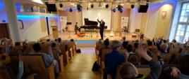 zdjęcie przedstawia prowadzącego koncert na scenie sali koncertowej podczas zabawy z uczestnikami koncertu siedzącymi na widowni, przy fortepianie akompaniator, na scenie instrumenty oraz banery szkoły i w tle ekran multimedialny z plakatem koncertu 