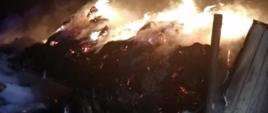 Płonące baloty ze słomą na zniszczonej prze ogień przyczepie rolniczej zlokalizowanej na polu ornym. Jest pora nocna.