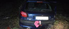 Kolizja samochodu osobowego. Na poboczu drogi polnej stoi samochód osobowy uczestniczący w zdarzeniu. Widok z zewnątrz od tyłu. Otwarte są drzwi auta z prawej strony. Na karoserii widoczne są wgniecenia, a także uszkodzona jest tylna lampa z prawej strony. Spod klapy bagażnika wystaje szmatka koloru różowego oraz trójkąt ostrzegawczy.