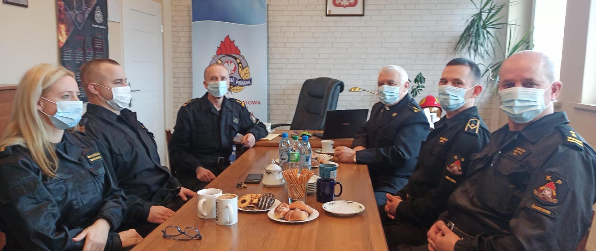 Zdjęcie przedstawia biuro Komendanta Powiatowego podczas warsztatów z Kapelanem zachodniopomorskich strażaków. Przy stole siedzi Kapelan oraz strażacy w tym jedna kobieta. Na stole znajdują się kubki oraz ciastka i paluszki.