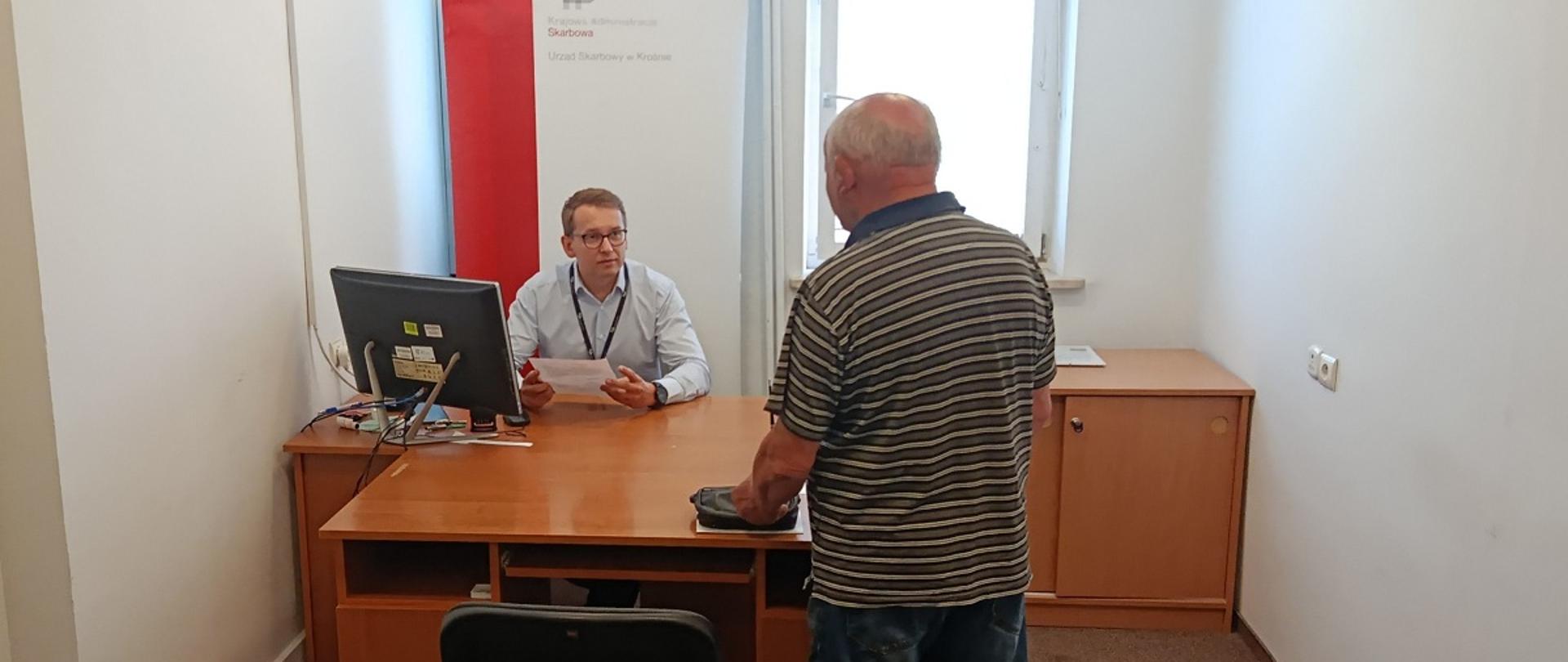 Przy biurku siedzi ekspert KAS. Obok stoi mężczyzna. W tle baner. 