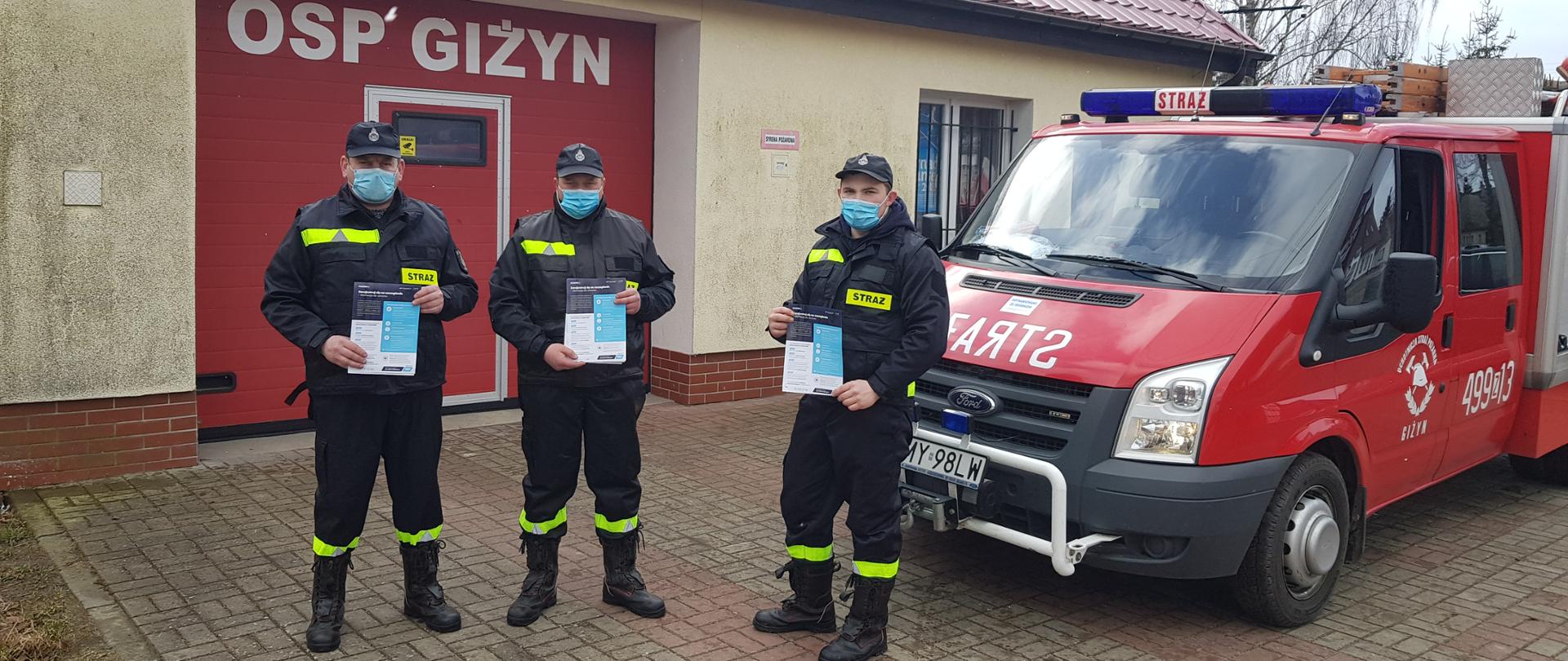 Druhowie ubrani w umundurowanie bojowe, trzymający ulotki w rękach stoją na tle samochodu pożarniczego i remizy strażackiej.