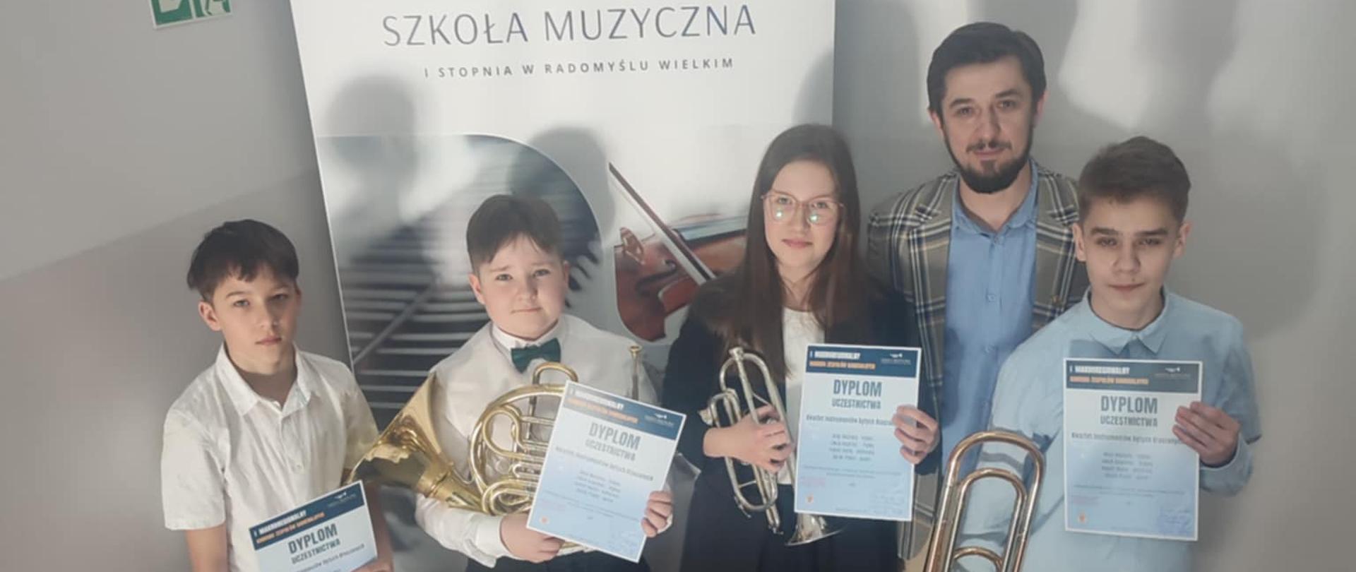 Na zdjęciu czworo młodych muzyków wraz z nauczycielem pozuje na klatce schodowej, trzymając w rękach dyplomy oraz instrumenty dęte blaszane: trąbki, waltornię i puzon. Za nimi stoi baner Szkoły Muzycznej I stopnia w Radomyślu Wielkim z grafiką trąbki i fortepianu. Wszyscy są elegancko ubrani i uśmiechają się, wyrażając dumę ze swojego osiągnięcia. Światło pada z góry, rzucając cienie na ścianę. Atmosfera zdjęcia podkreśla radość i prestiż konkursowego sukcesu.