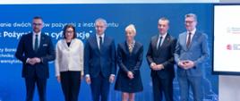 Przed wielkim niebieskim ekranem z napisem KPO: pożyczka na cyfryzację stoją: minister Kulasek, kobieta w białej marynarce, mężczyzna w garniturze, kobieta w granatowej marynarce i dwóch mężczyzn w garniturach.