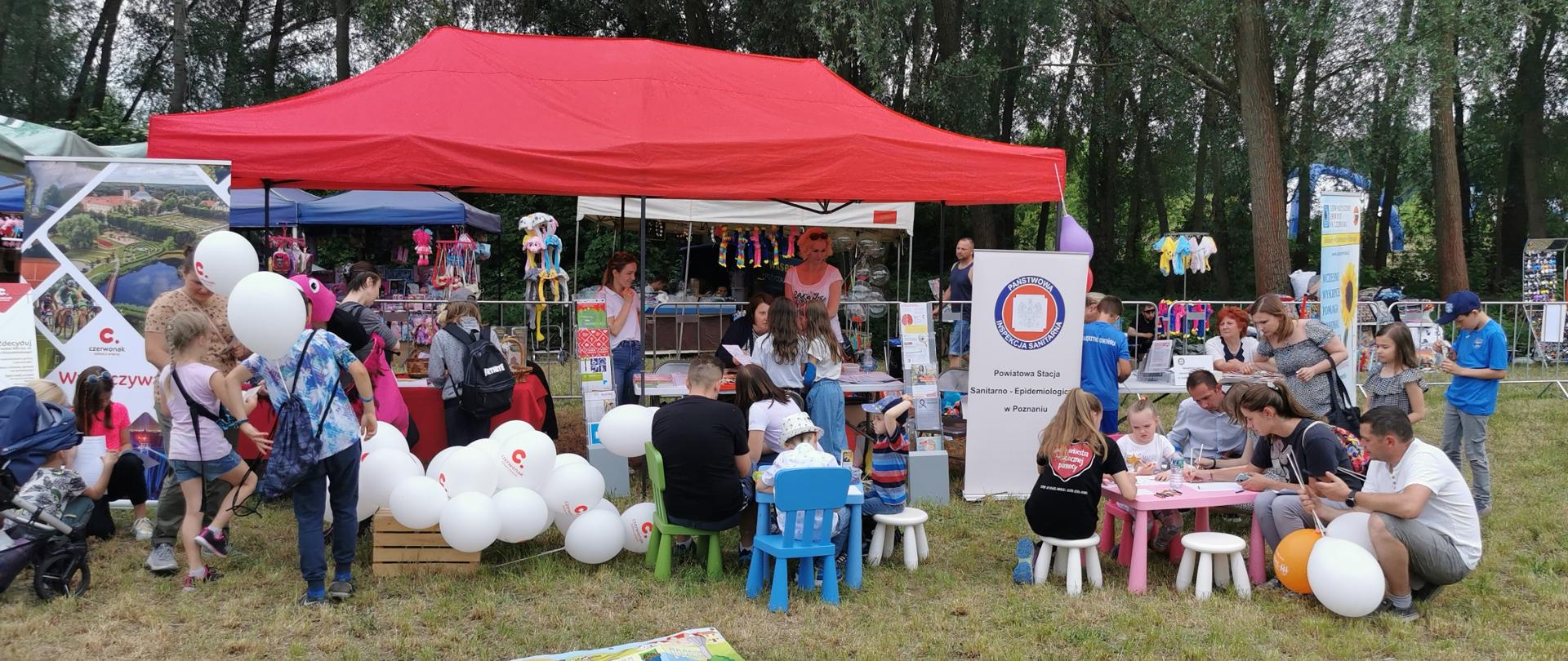 Stanowisko PSSE w Poznaniu oraz gminy Czerwonak. Przed namiotem przy stolikach dzieci i dorośli rozwiązują zadania konkursowe.