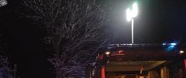 Na zdjęciu widzimy samochód pożarniczy podczas działań przed budynkiem objętym pożarem. Zdjęcie wykonane w porze nocnej.