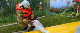 na 1 planie strażak trzyma węża do napełnienia rękawów przeciwpowodziowych, dalej strażak w wodzie trzymający się linki, na brzegu strażak go asekuruje linką