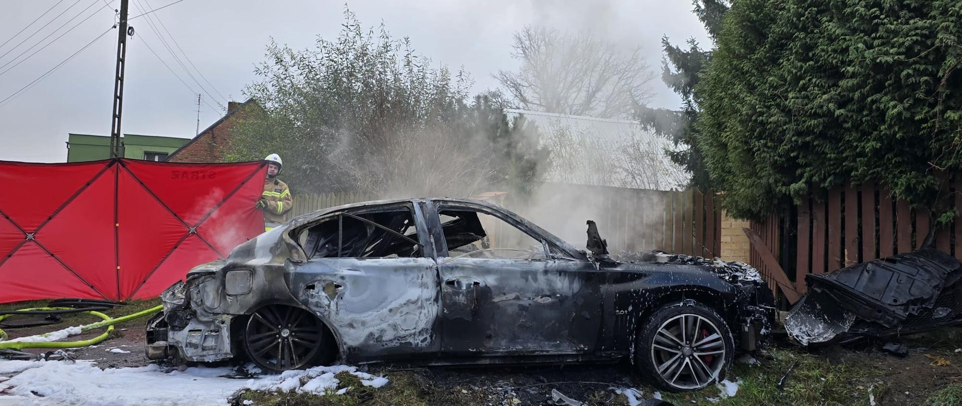 Spalony samochód na poboczu przy płocie, za nim czerwony parawan i strażak
