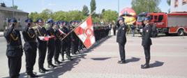 Na zdjęciu widzimy strażaków stojących w szeregu. Strażacy ubrani są w galowe mundury koloru czarnego. Jeden ze strażaków trzyma sztandar opuszczony w dół . Przed strażakami stoi przodem kolejnych dwóch strażaków i oddaje w ich kierunku honory w postaci zasalutowania. W tle widzimy budynki mieszkalne, zielone drzewa a także wóz strażacki koloru czerwonego.