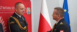 Świetlica KW PSP w Poznaniu. Wielkopolski Komendant Wojewódzki PSP wręcza funkcjonariuszowi PSP decyzję o powierzeniu obowiązków na stanowsiku komendanta powiatowego PSP w Rawiczu. Funkcjonariusze ściskaja sobie prawe dłonie. W tle ścianka promocyjna PSP oraz flagi RP i UE