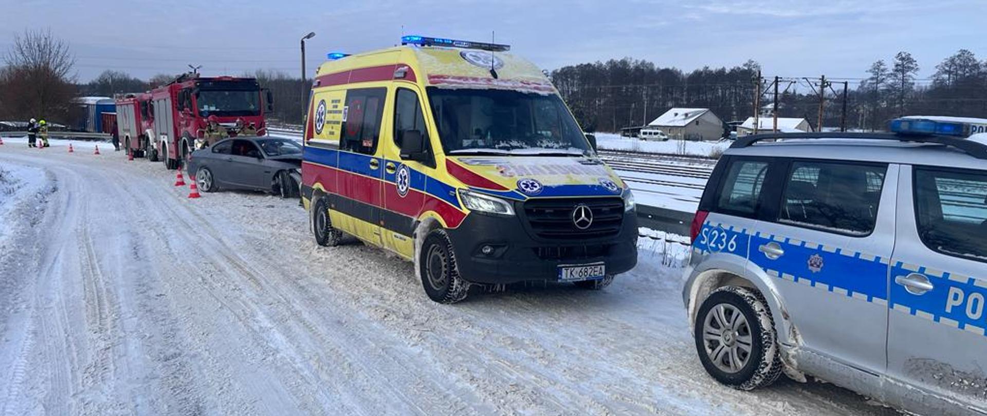 Na zdjęciu widać dwa samochody pożarnicze, karetkę pogotowia oraz radiowóz policyjny. Samochody zaparkowane na zaśnieżonej drodze. W środku, między nimi, znajduje się uszkodzony samochód po wcześniejszej kolizji.