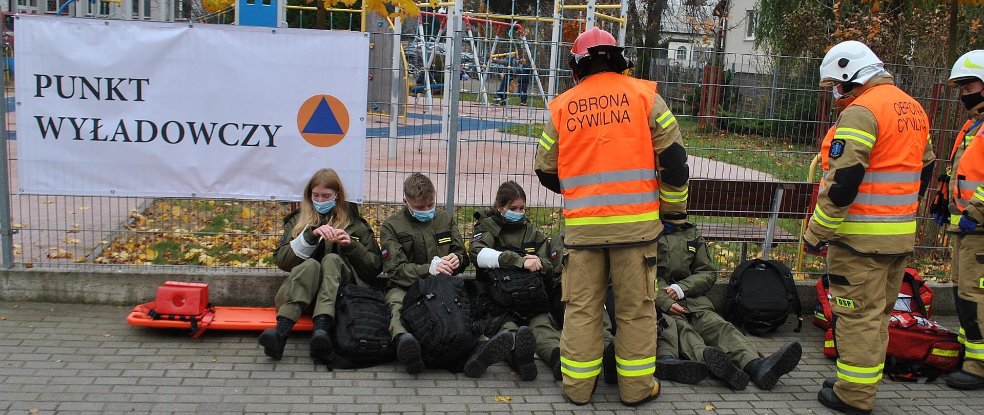 Na zdjęciu znajdują się strażacy, udzielający pomocy ewakuowanym w punkcie wyładowczym. Ewakuowani to uczniowie, biorący udział w ćwiczeniu w charakterze pozorantów.