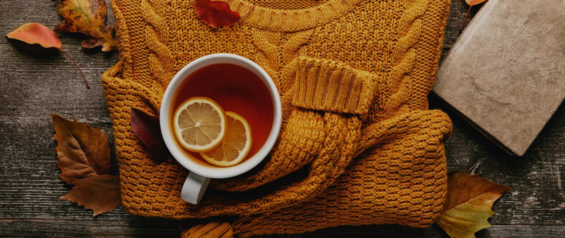 Na zdjęciu znajduje się sweter, książka i herbata z cytryną. W tle widoczne są jesienne liście.