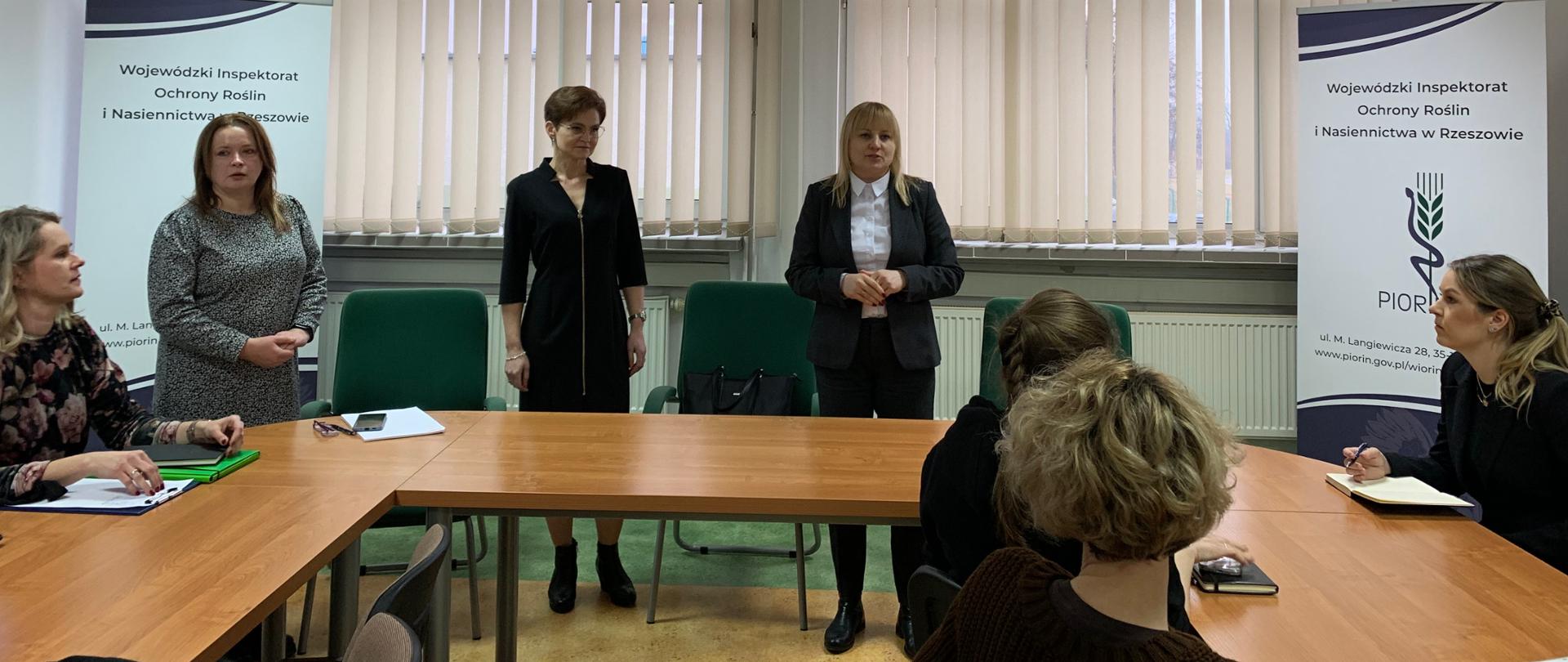 Trzy stojące kobiety, ubrane na elegancko. Kobieta po lewej ubrana jest w szarą sukienkę, kobieta na środku ubrana jest w czarną sukienkę a kobieta po prawej stronie ubrana jest w czarny garnitur i białą koszulę. W pomieszczeniu za kobietami znajduję się duże okno z jasnymi roletami. W pomieszczeniu panują jasne kolory. Po prawej i lewej stronie stoją dwa duże roll-upy z logiem instytucji.
W pomieszczeniu znajdują się stoły przy których siedzi kilka osób.
