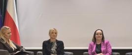 Zdjęcie przedstawia scenę z panelu dyskusyjnego, trzy kobiety siedzą w fotelach ustawionych w rzędzie. Kobieta w środku, blondynka, trzyma mikrofon i mówi do niego. W tle, za uczestniczkami, widać dwie flagi: jedną z gwiazdami na niebieskim tle oraz drugą w biało czerwonych – barwach.
