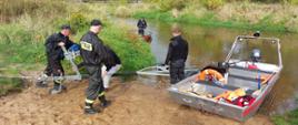 strażacy na brzegu rzeki przy łodzi strażackiej podczas wodowania łódki, w tle drzewa i krzewy