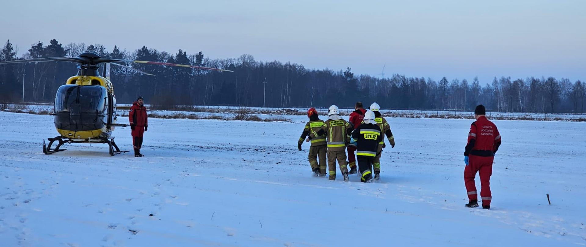 Helikopter Lotniczego Pogotowia Ratunkowego stoi na polu na śniegu. Strażacy z ratownikami niosą na noszach poszkodowaną kobietę