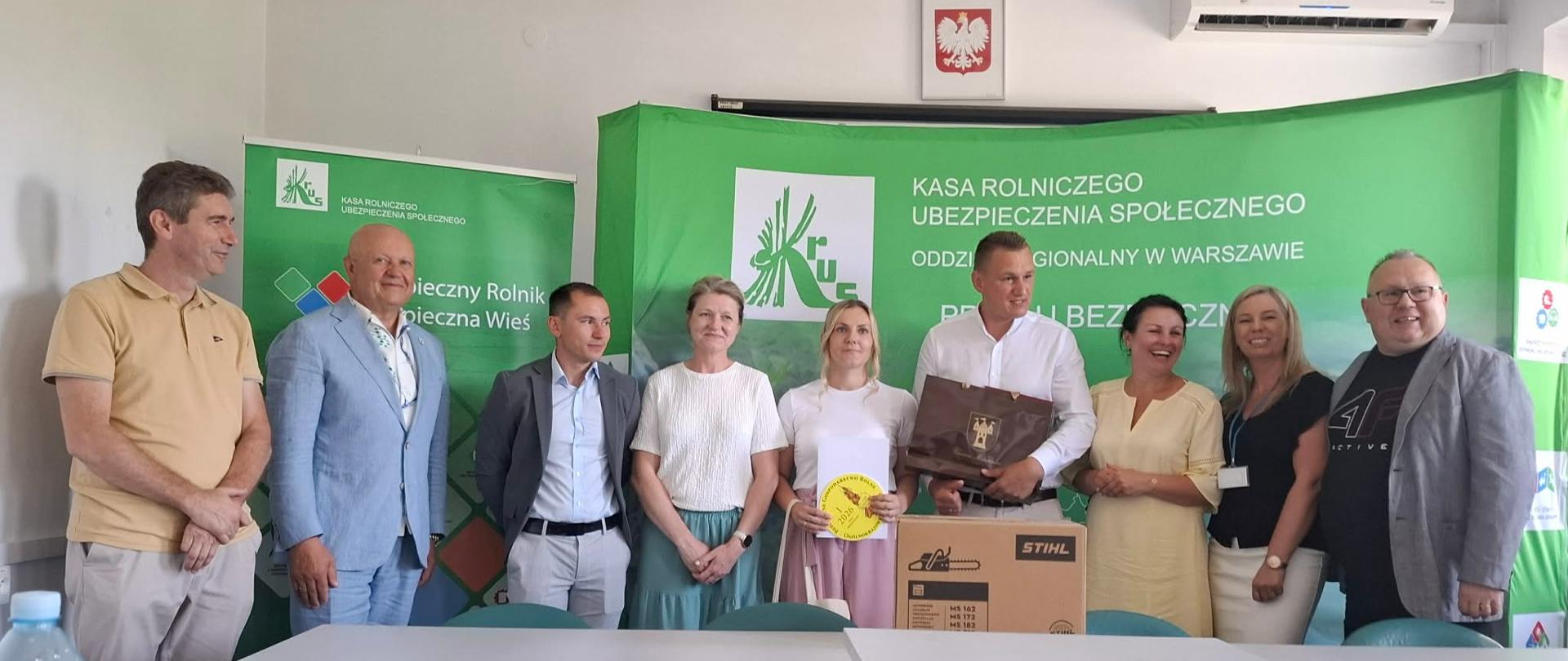 W pomieszczeniu znajduje się dziewięć osób. pięciu mężczyzn oraz cztery kobiety. Jeden z mężczyzn trzyma w ręku torebkę z nagrodą natomiast jedna z kobiet trzyma w ręku medal i dyplom za zajęcie I miejsca w konkursie. Na stole leży główna nagroda - pilarka.