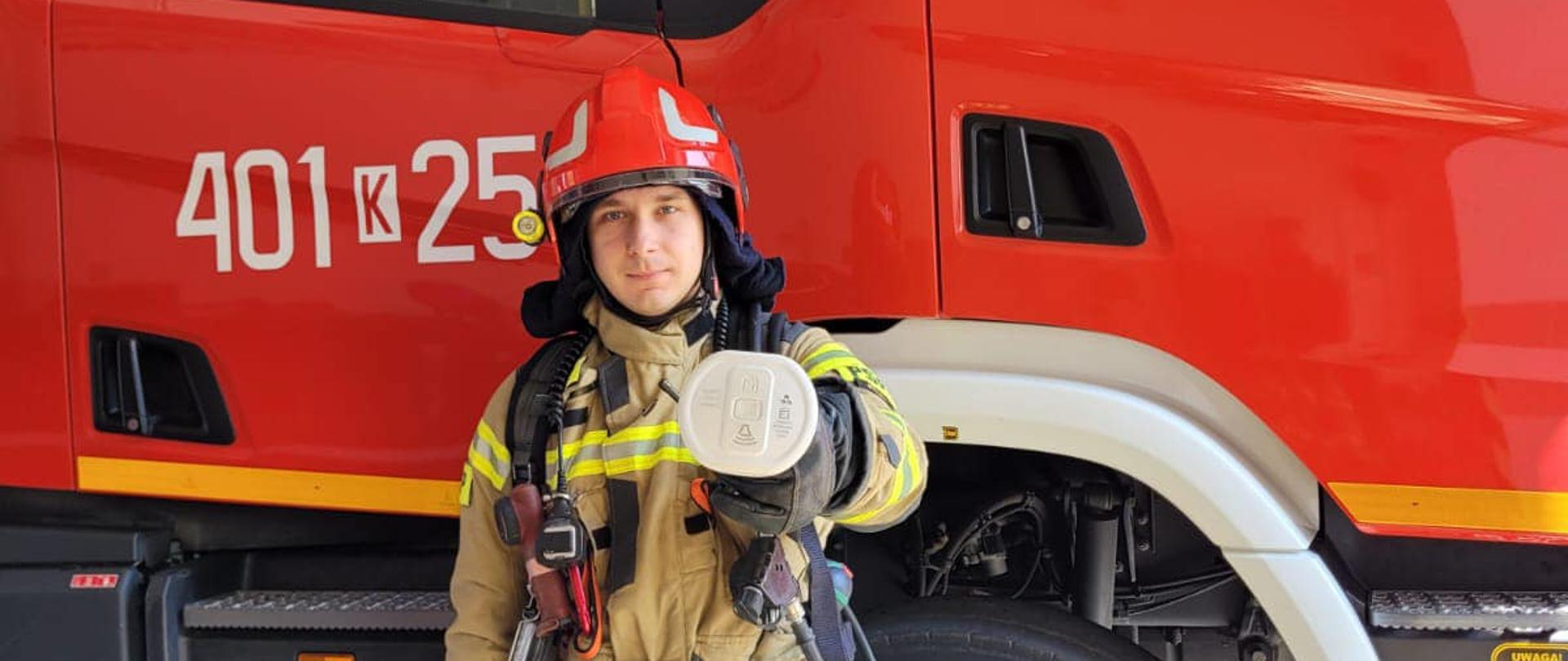 Na zdjęciu znajduje się strażak ubrany w ubranie specjalne i hełm trzymający czujkę dymu w ręce. W ple znajduje się duży czerwony samochód strażacki. 