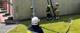 druhowie w trakcie egzaminu epizodu z zakresu pożarów dachów ubrani w aparatu ochrony dróg oddechowych, czarne i piaskowe ubrania specjalne do dachu budynku dostawiona jest drabina na której stoi strażak.