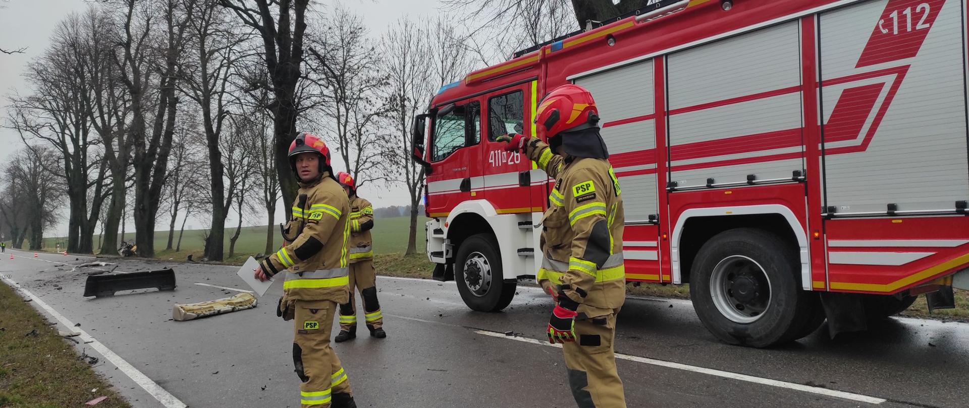 Na drodze stoi trzech strażaków w czerwonych hełmach. Z prawej strony duży czerwony samochód strażaki. Na drodze z lewej porozrzucane elementy pojazdu. Pochmurnie.