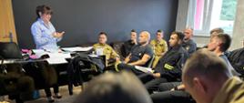 Grupa około 20. strażaków siedzi i słucha wykładu. Prowadzącą kobieta. Strażak z prawej strony notuje.
