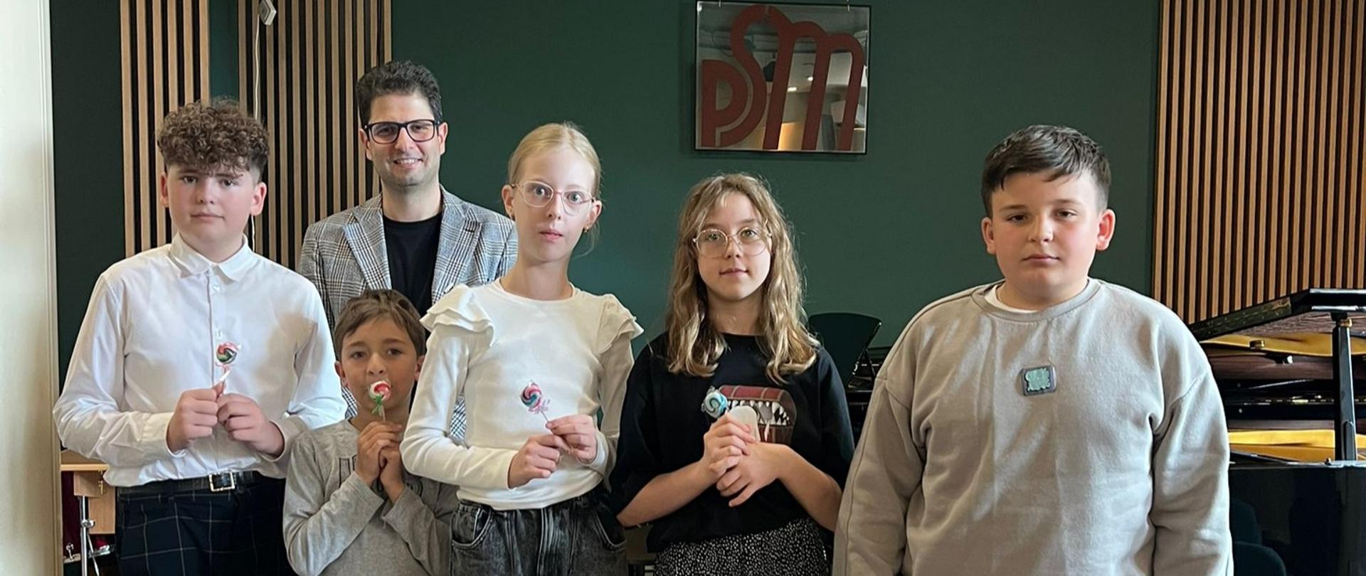 Pięcioro uczniów ustawionych w rzędzie z mężczyzną w marynarce stojącym z tyłu; dzieci trzymają lizaki, za nimi fortepian i zielona ściana z logo — zdjęcie grupowe po występie w szkole muzycznej.