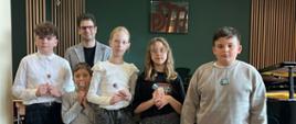Pięcioro uczniów ustawionych w rzędzie z mężczyzną w marynarce stojącym z tyłu; dzieci trzymają lizaki, za nimi fortepian i zielona ściana z logo — zdjęcie grupowe po występie w szkole muzycznej.