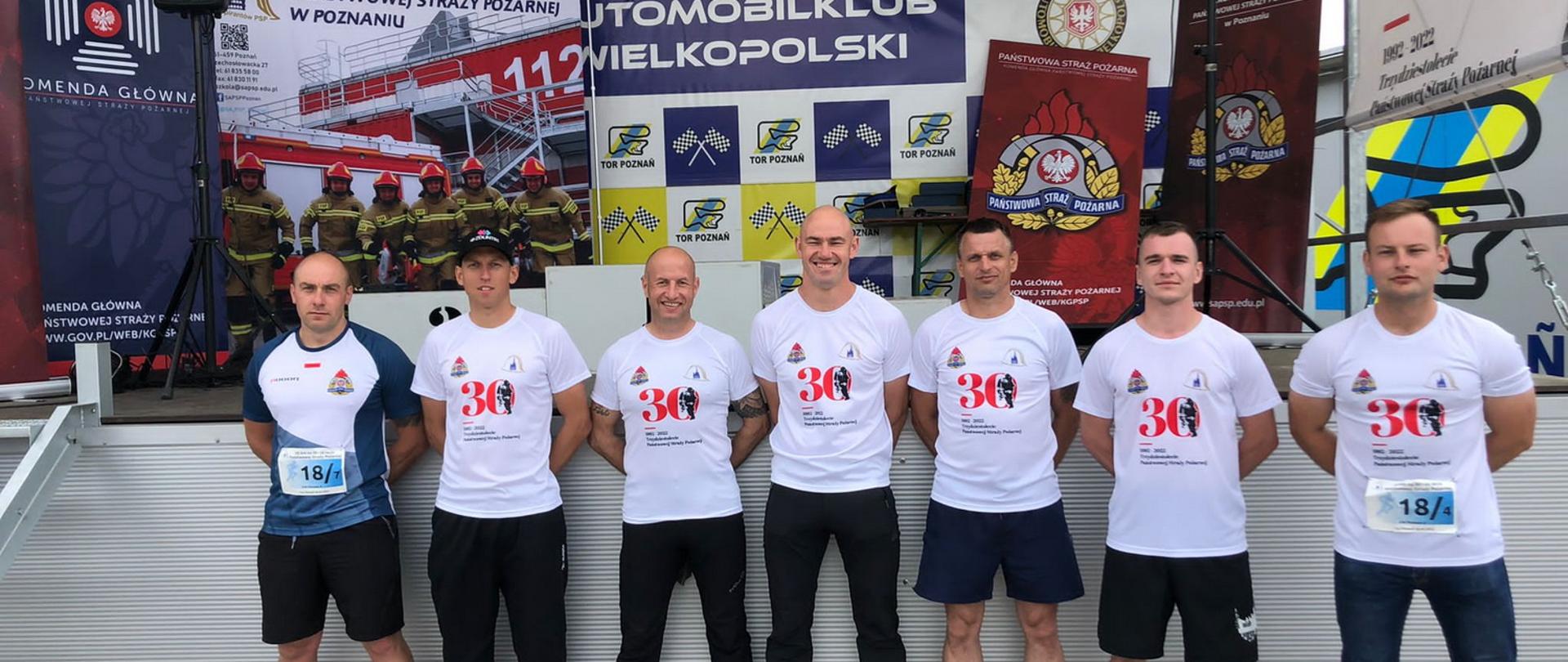 Grupa siedmiu mężczyzn w strojach sportowych stoi przed saceną na której ustawione są banery