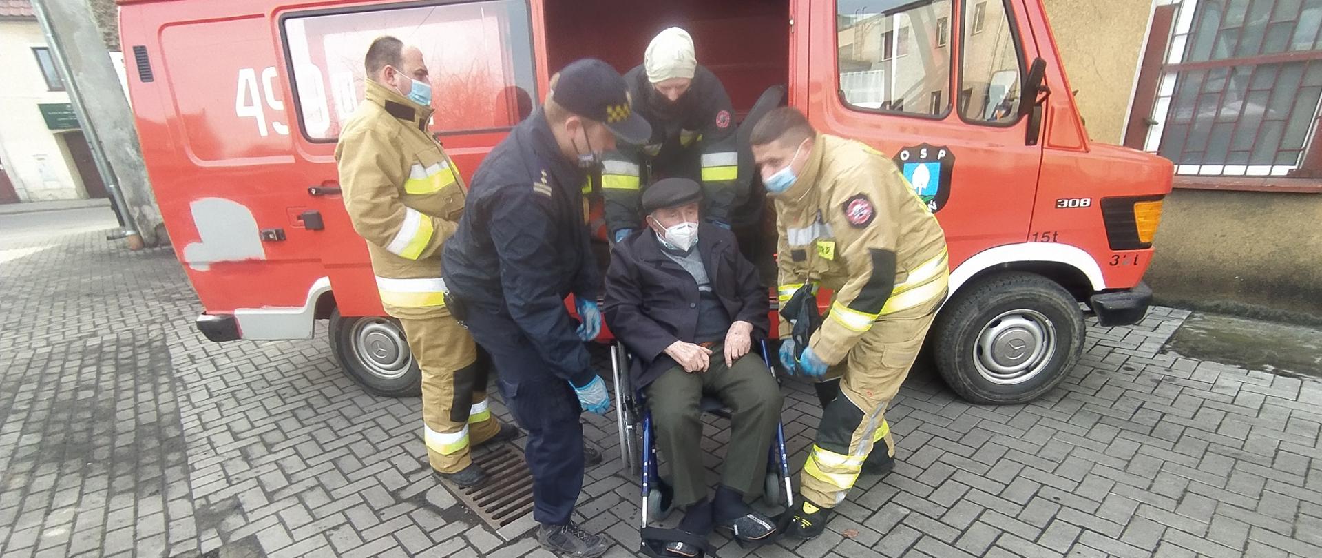 Zdjęcie przedstawia trzech strażaków z jednostki OSP Wleń oraz strażnika miejskiego, którzy pomagają wsiąść do samochodu starszemu mężczyźnie podczas transportu na szczepienie przeciwko Covid-19. 