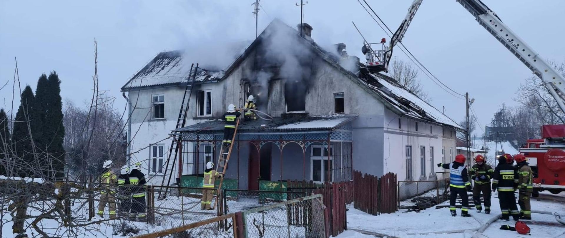 Strażacy podczas gaszenia pożaru piętrowego budynku mieszkalnego. Z okien wydobywa się dym. Strażak stojący na drabinie trzyma linię gaśniczą, obok na daszku znajduje się drugi strażak, który kieruje prąd wody do pomieszczenia przez okno na pierwszym piętrze. Po prawej stronie stoi podnośnik hydrauliczny. Obok stoją strażacy.