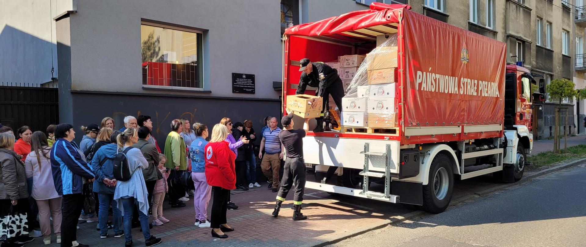 Na zdjęciu samochód pożarniczy kontener. Przed nim grupa uchodźców wojennych którym kadeci wydają dary