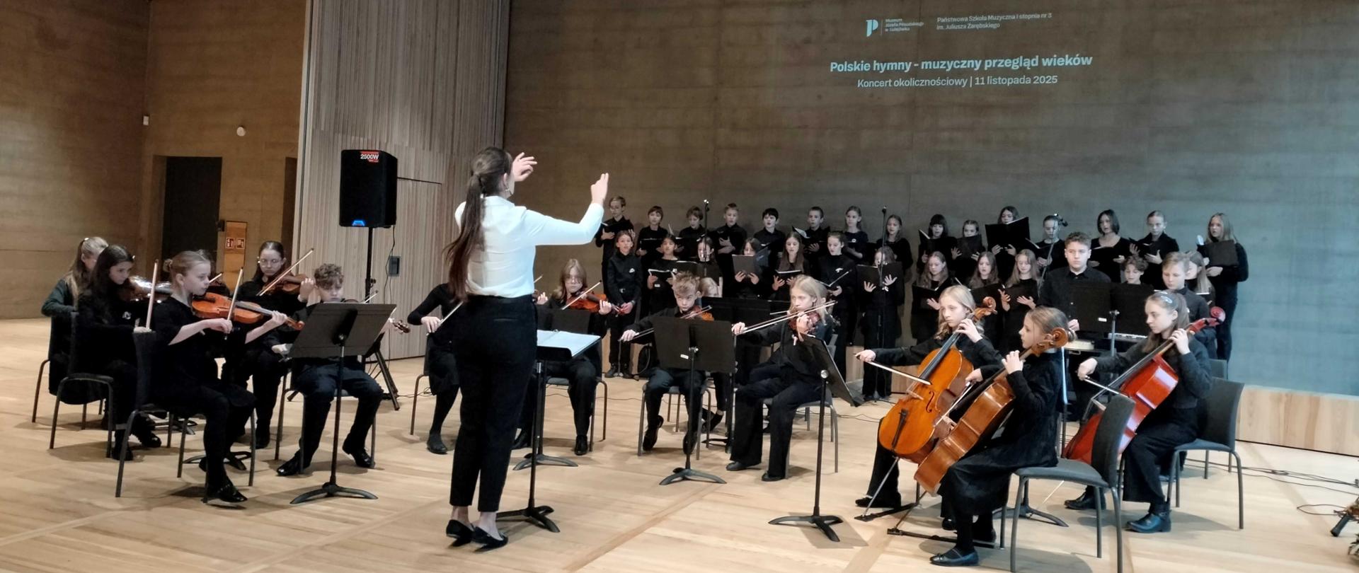 chór i orkiestra uczniów gra i śpiewa na scenie pod dyrekcją nauczycielki