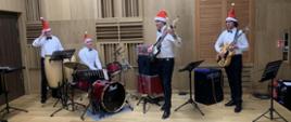 Na scenie widocznych jst czterech mężczyzn. Każdy z nich ma na głowie czerwoną czapkę Mikołaja. Patrzac od lewej dwaj mężczyźni grają na instrumentach perkusyjnych, kolejni dwaj grają na gitarach.