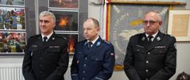 Na zdjęciu podczas wernisażu wystawy „Na STRAŻY bezpieczeństwa mieszkańców Ziemi Gorlickiej" widoczni w ubraniach galowych koloru granatowego po lewej stronie zastępca komendanta powiatowego Państwowej Straży Pożarnej w Gorlicach w środku komendant powiatowy policji a po prawej stronie wiceprezes zarządu powiatowego Związku Ochotniczych Straży Pożarnych RP w Gorlicach. W tle widoczne zdjęcia i eksponaty pożarnicze sztandar, hełm, ubranie strażackie typu moro w gablocie.