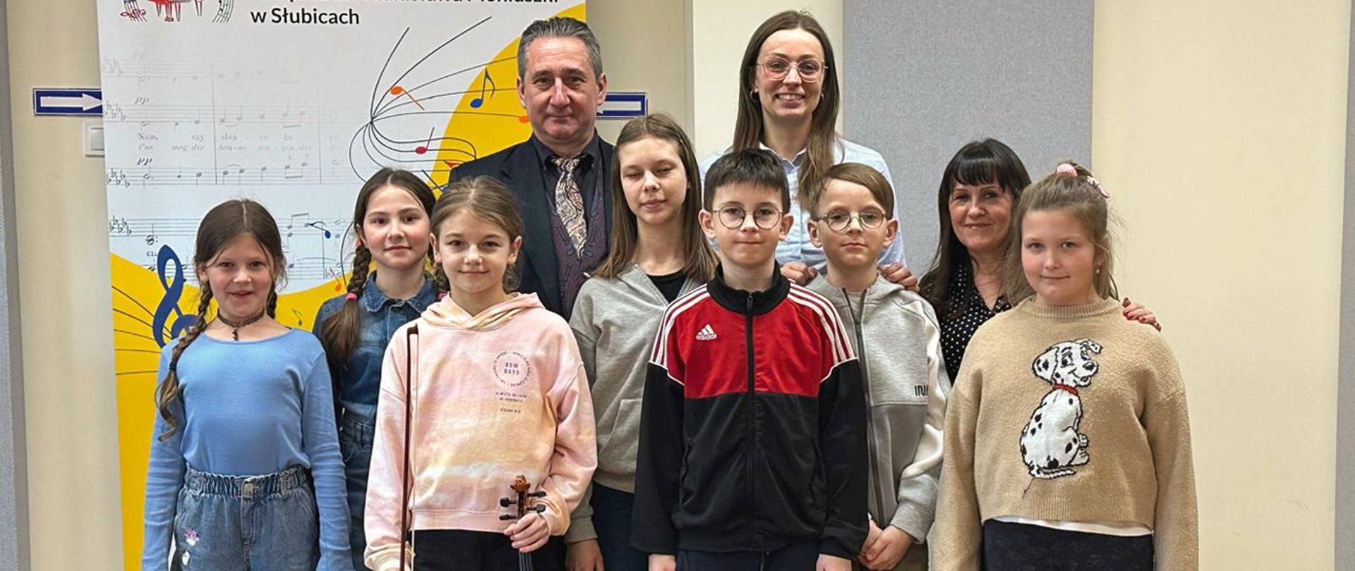 Na zdjęciu grupa osób, troje dorosłych i siedmioro dzieci stoją w auli szkolnej na tle baneru szkoły.