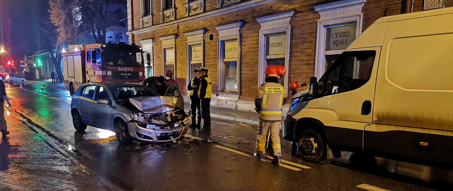 Na zdjęciu widoczne dwa pojazdy po zderzeniu przód samochodu osobowego i lewy bok samochodu dostawczego. Komora silnika częściowo uszkodzona po lekkim zderzeniu czołowym. Natomiast na zdjęciu nie widać przodu samochodu dostawczego. Przed samochodami stoją strażacy , a w oddali policjanci i wóz strażacki.