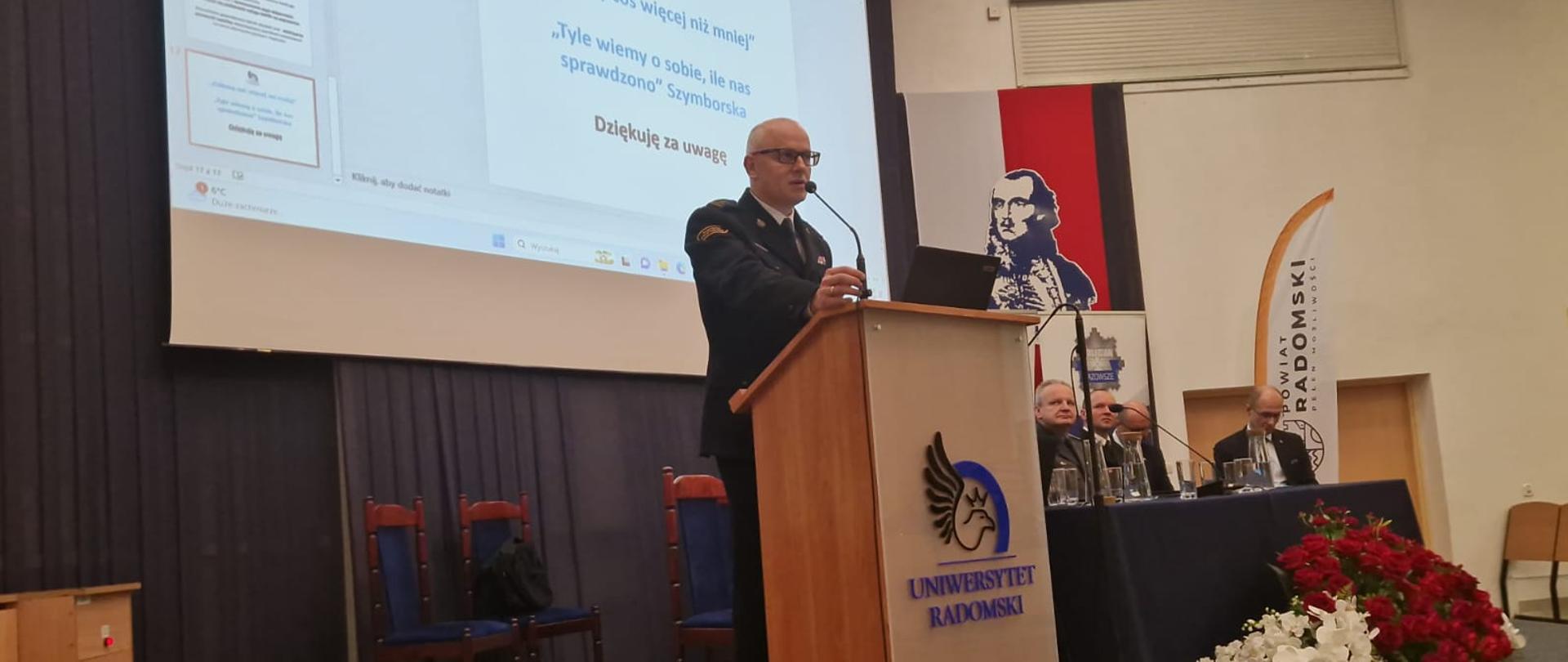 Zdjęcie wykonane podczas konferencji naukowej poświęconej tematyce bezpieczeństwa w regionie radomskim. Konferencja zrealizowana na auli uniwersytetu humanistycznego w Radomiu. Na zdjęciu od lewej widać Zastępcę Komendanta Głównego Państwowej Straży Pożarnej starszego brygadiera Pawła Frysztaka. Funkcjonariusz stoi na mównicy skąd wygłasza swoją prelekcję dotyczącą bezpieczeństwa. W oddali po prawej widać ustawiony stół przy którym siedzą inni prelegenci uczestniczący w konferencji. 