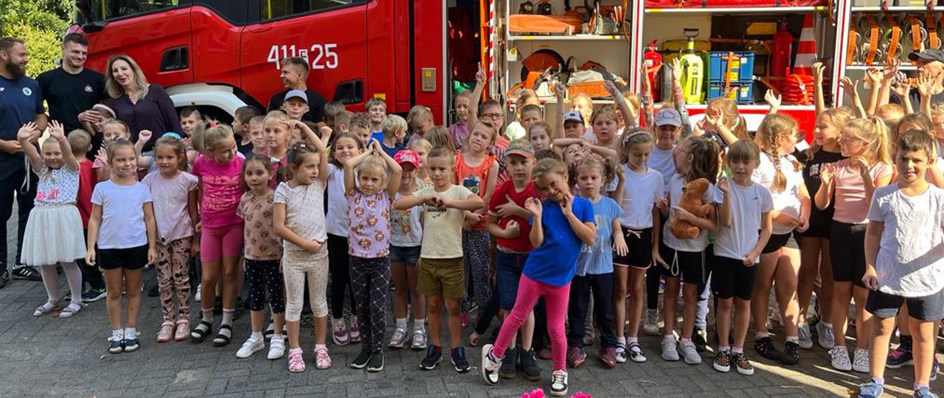 Jest to zdjęcie grupowe dzieci, wychowawców i strażaków na tle samochodu gaśniczego z otwartymi skrytkami sprzętowymi.