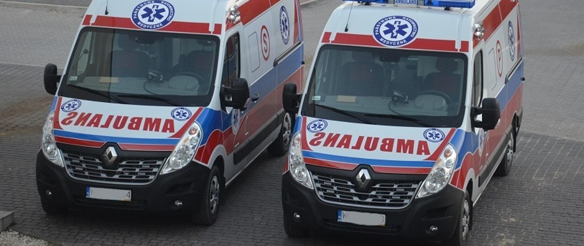 Dwa samochody, stojące obok siebie, służące do transportu pacjentów. Oznakowane słowem ambulans i logiem Państwowego Ratownictwa Medycznego. 