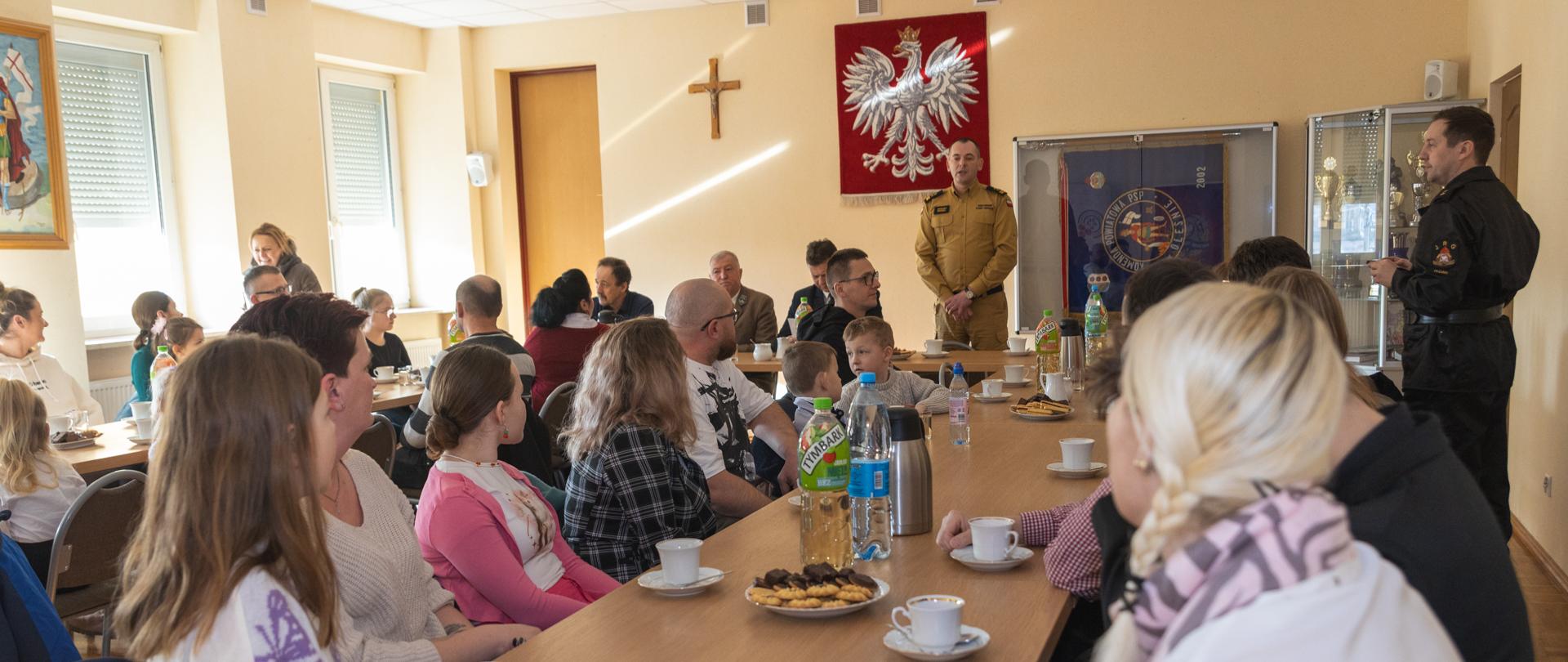 Uczestnicy spotkania siedzą przy stołach w sali konferencyjnej Komendy Powiatowej Państwowej Straży Pożarnej w Oleśnie. W centralnym miejscu stoi Komendant Powiatowy PSP w Oleśnie - bryg. Andrzej Wróbel, który wita przybyłe osoby. W tle znajduje się Godło Polski.