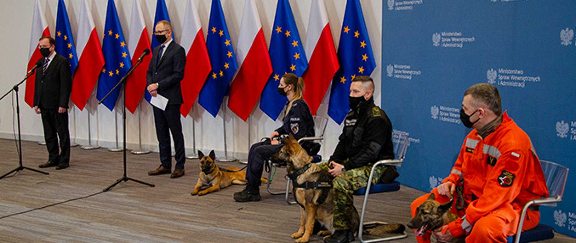 na zdjęciu przedstawiona jest konferencja prasowa ministra mariusza kamińskiego, który stoi na tle biało-czerwonych i unijnych flag, na krzesłach siedzą opiekunowie psów służbowych Policji, Straży Granicznej i Państwowej Straży Pożarnej wraz ze swymi podopiecznymi.